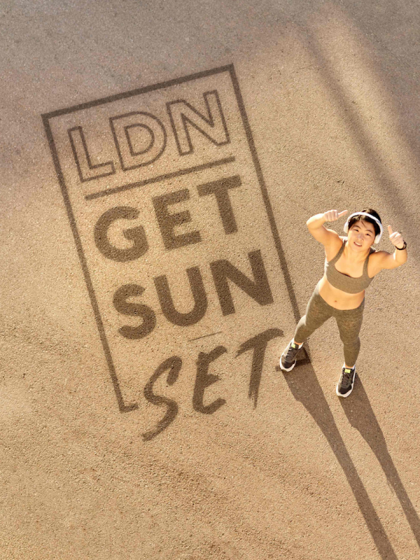 A runner is standing on sand giving the thumbs up to the sky. On the sand are the words LDN Get Sun Set.