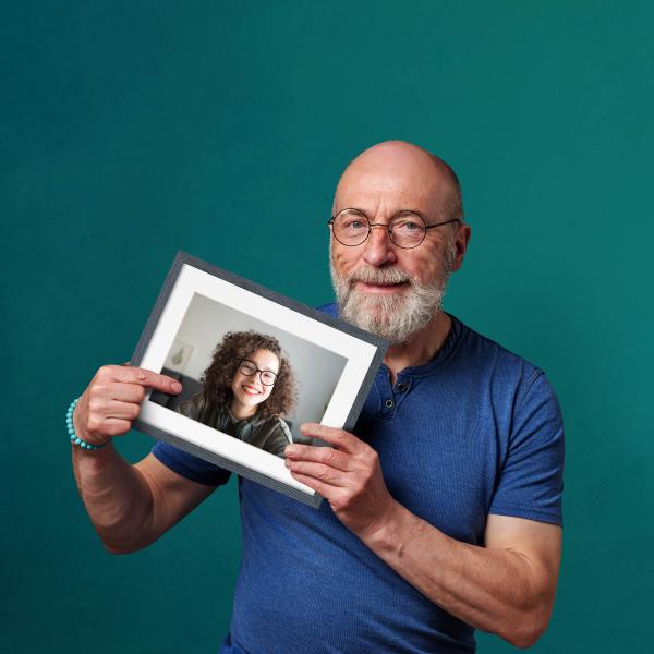 A man with a white beard is holding up a picture of a younger female family member.